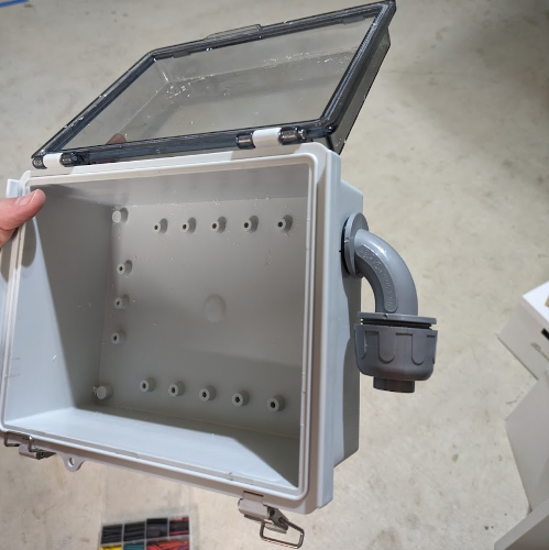Empty waterproof junction box with a clear lid and an elbow conduit connector sticking out of the side